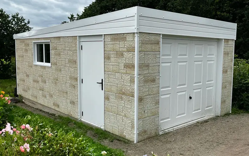 STONE-CLAD-GARAGE - All Seasons Steel Buildings Stone outbuilding with beige brick pattern, white side door, small window, and large white garage door on a dirt path.