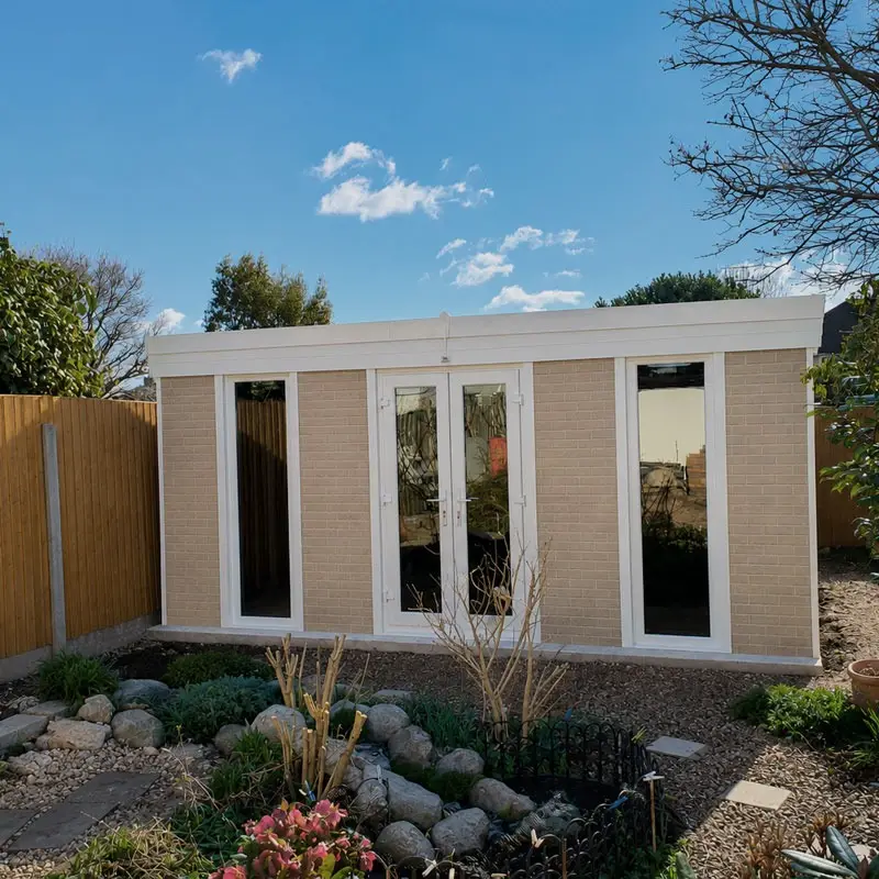 Small beige brick garden room with white-framed glass doors in a fenced backyard with a rock garden and plants.