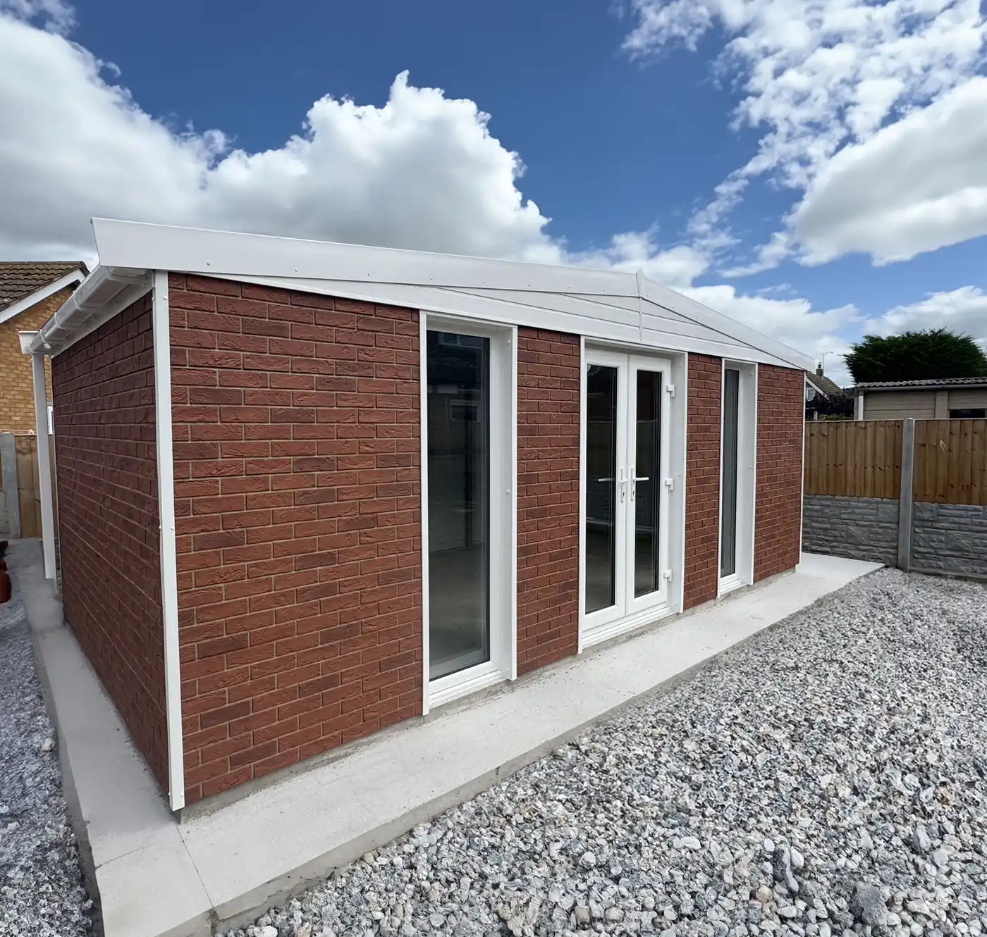 New brick extension with white-framed glass doors, set on a concrete foundation, surrounded by gravel yard under a blue sky with clouds.