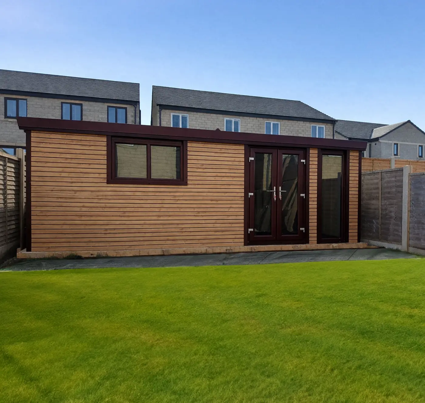 Wooden garden shed in a backyard with a green lawn and brick houses against a clear blue sky.
