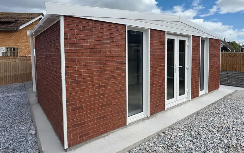 BRICK-CLAD-GARDENROOM - All Seasons Steel Buildings Red brick extension with white-framed glass doors along a concrete patio, surrounded by a gravel yard and wooden fence.