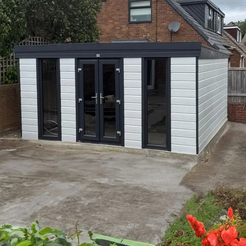 Modern gray garden room with double glass doors and tall side window, on a concrete patio beside a brick house, with red flowers in the foreground.