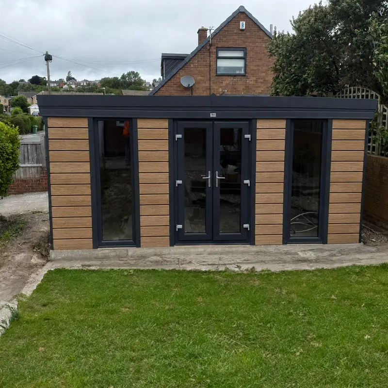 Garden room or modern shed with dark frame and wooden paneling, glass doors, and large side windows in a backyard setting.